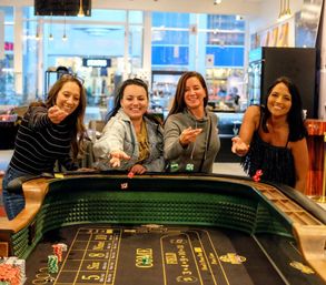 Four friends smiling and throwing dice at a casino craps table on a lively indoor gaming floor with chips and onlookers in the background.