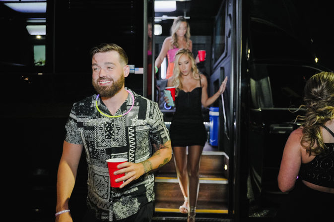 Smiling bearded man in a patterned shirt holding a red cup exits a party bus at night while two women in cocktail dresses follow down illuminated steps, neon glow necklaces adding a festive nightlife vibe.