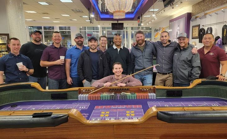 Group of smiling men posing behind a purple-felt craps table in a casino setting, with stacked chips, a dealer’s stick and a sparkling chandelier overhead.