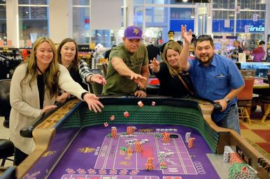 Five people leaning over a purple craps table, laughing and tossing dice on a lively indoor casino gaming floor.