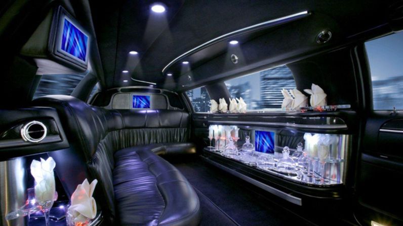 Luxury stretch limousine interior with curved black leather bench seats, illuminated glassware bar, blue LED ambient lighting and blurred city skyline visible through windows