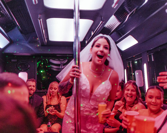 Bridal party on a party bus: laughing bride in a wedding dress and veil holding a dance pole and a drink, surrounded by guests with cups under colorful disco lights.