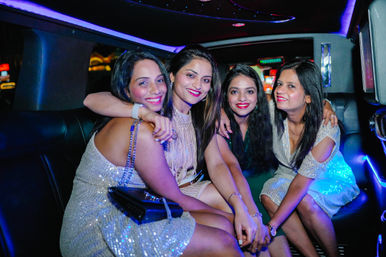 Four friends in sparkly dresses smiling and posing together inside a stretch limousine with colorful LED lights during a nighttime celebration