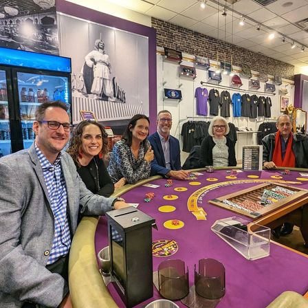 Six adults smiling around a purple-felt poker table with chips and cards in a casino-style indoor gaming lounge and retail shop, merchandise on the wall and a large black-and-white mural in the background.