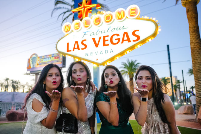 Four women in party dresses blowing kisses at the camera in front of the illuminated 'Welcome to Fabulous Las Vegas, Nevada' sign at dusk