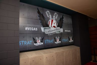 Indoor wall display at a Las Vegas comedy club featuring a winged microphone logo, multiple comedy badges and ‘#VEGAS’ hashtags.