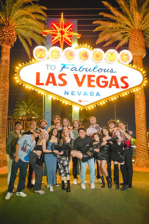 Large group of friends posing at night beneath the glowing Welcome to Fabulous Las Vegas, Nevada sign, palm trees and neon lights creating a festive party backdrop.
