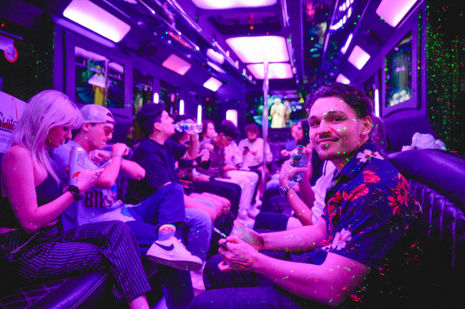 Group of young adults partying inside a neon-lit party bus interior with purple LED panels and green laser dots, seated on leather benches and holding drinks in a lively nightlife scene