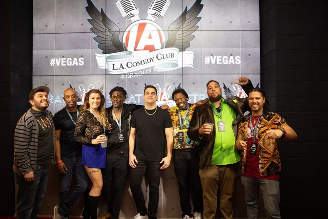 Eight people posing and smiling in front of a comedy-club backdrop with winged microphone logo and #VEGAS signage at a Las Vegas comedy night.
