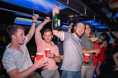 Friends partying on a neon-lit party bus, holding red cups and a raised champagne bottle under blue LED lights