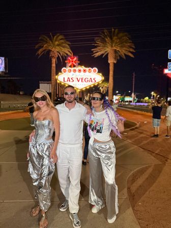Three people posing at night in front of the iconic Welcome to Fabulous Las Vegas sign — two women in shiny silver outfits and a man in white, all wearing sunglasses with palm trees and neon lights in the background.