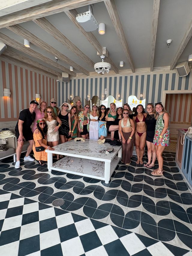 Group of friends in swimwear and summer outfits posing in a striped coastal lounge at a beachside resort, with patterned black-and-white tile floor, marble coffee table and wooden beamed ceiling — cheerful vacation gathering.