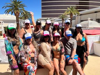 Cheerful daytime pool party: a diverse group in colorful swimwear and novelty trucker hats posing and smiling on a sunny resort pool deck with palm trees and a tall hotel tower in the background.