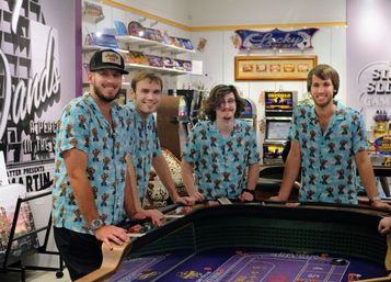 Four young men in matching blue tropical shirts smiling around a casino craps table with chips and slot machines in the background.
