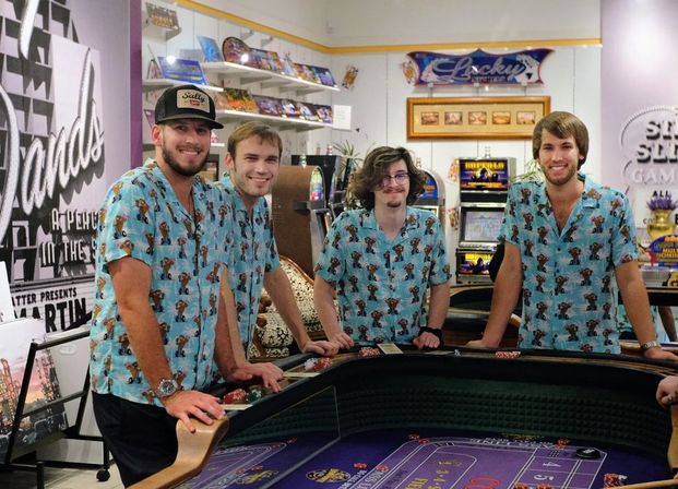 Four young men in matching blue tropical shirts smiling around a casino craps table with chips and slot machines in the background.