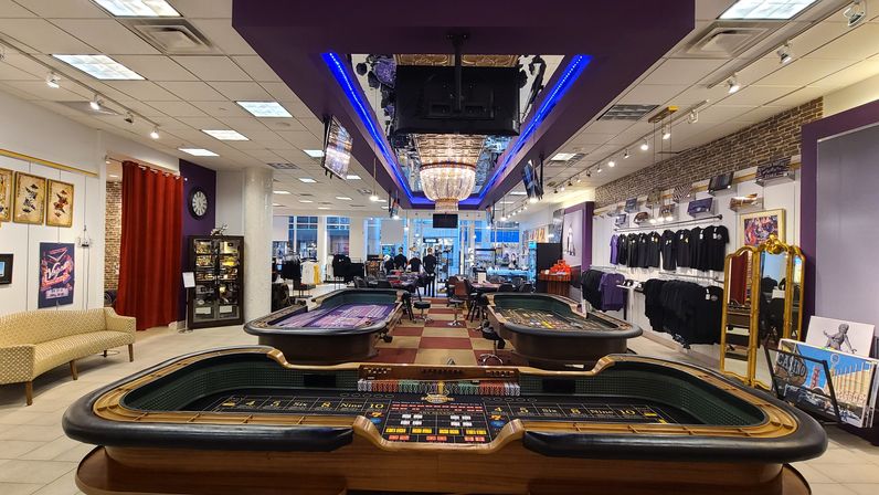 Urban storefront showroom with casino-style gaming tables in foreground, illuminated chandelier and blue-purple ceiling trim, apparel racks, displays and lounge seating in a bright retail space