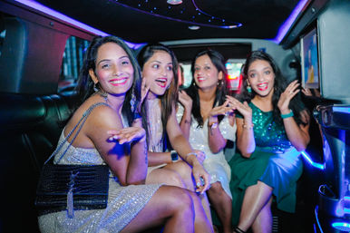 Four smiling women in sparkly dresses celebrating inside a luxury stretch limousine with neon lights, posing for a fun night-out in the city.