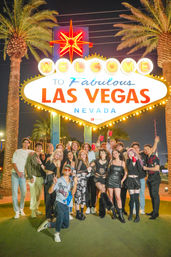 Group of friends posing at night beneath the neon "Welcome to Fabulous Las Vegas Nevada" sign with palm trees, glowing lights and a festive party vibe
