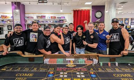 Group of eleven people in casual matching shirts smiling and posing behind a casino-style craps table loaded with colorful poker chips in a bright indoor gaming room with game shelves and a wall clock.