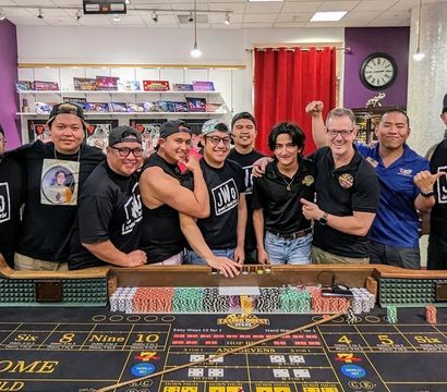 Group of eleven people in casual matching shirts smiling and posing behind a casino-style craps table loaded with colorful poker chips in a bright indoor gaming room with game shelves and a wall clock.