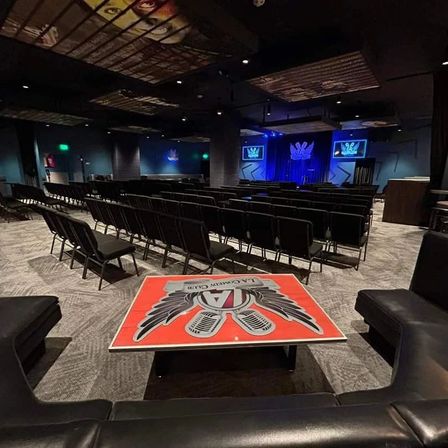 Modern indoor comedy club or event venue with rows of black chairs facing a blue-lit stage, twin screens on either side, and a red logo-topped table in the foreground.