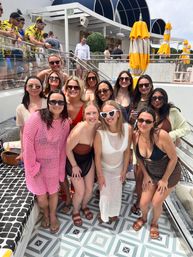 Group of smiling women at a sunny outdoor pool club day party, posing on geometric tiles in swimsuits and cover-ups with sunglasses, yellow umbrellas and lounge seating in the background.
