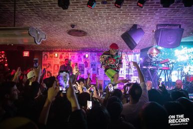 Live concert in an intimate speakeasy-style club with brick ceiling — energetic performer in colorful outfit and red beanie jumps on stage as a packed crowd holds up phones under purple-blue lights.