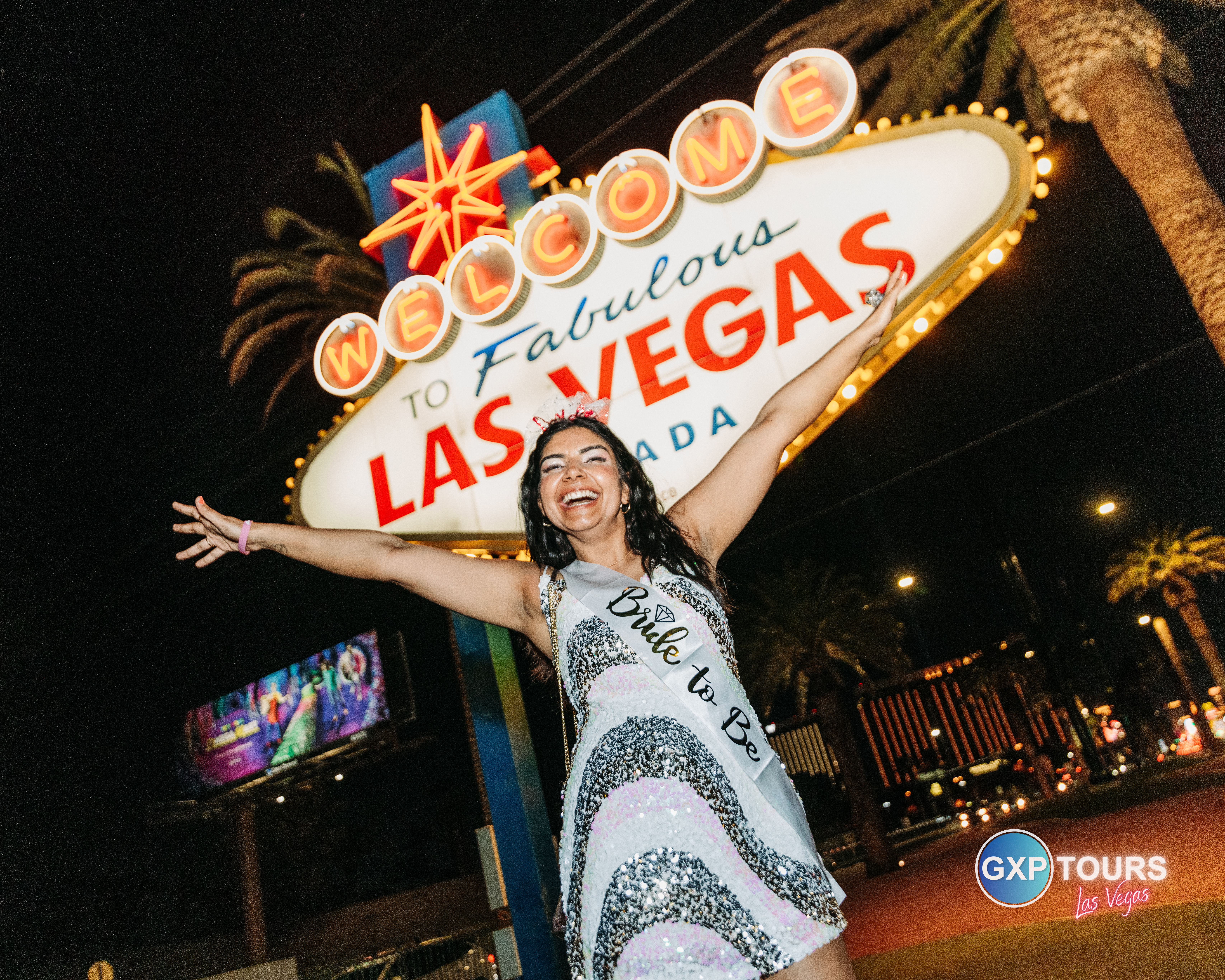 Person in a sequined dress wearing a Bride to Be sash and tiara, arms outstretched and smiling in front of the illuminated Welcome to Fabulous Las Vegas sign at night