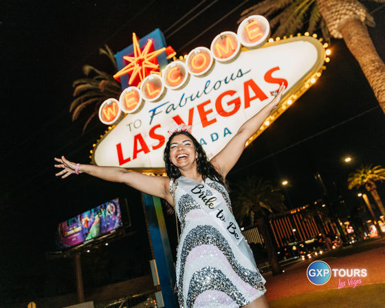 Person in a sequined dress wearing a Bride to Be sash and tiara, arms outstretched and smiling in front of the illuminated Welcome to Fabulous Las Vegas sign at night