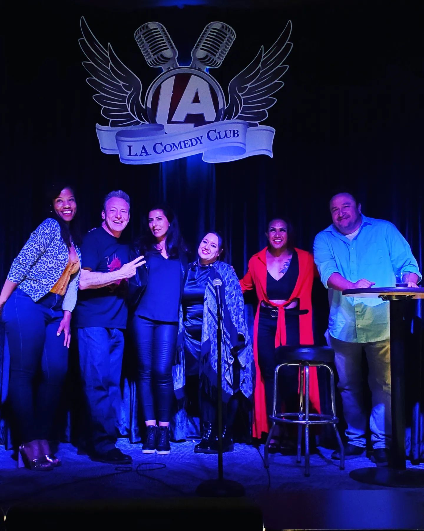 Six comedians smiling and posing on a Los Angeles comedy club stage beneath a microphone-and-wings logo, bathed in purple-blue stage lighting with a stool and mic stand nearby.