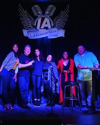 Six comedians smiling and posing on a Los Angeles comedy club stage beneath a microphone-and-wings logo, bathed in purple-blue stage lighting with a stool and mic stand nearby.