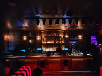 Dim urban cocktail bar interior with backlit wooden liquor shelves, tufted red leather bar front, overhead stage lights and bartenders serving guests in warm amber and red lighting.