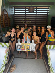 A lively birthday celebration in a shaded resort pool cabana — a dozen women in swimsuits cheer with shot glasses and oversized liquor bottle cutouts under a 'Happy Birthday' banner.