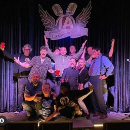 Group of comedians and friends posing on a Los Angeles comedy club stage under a winged microphone logo, laughing and cheering with red cups and a small dog, purple stage lighting and black curtains.
