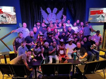 Large group smiling and posing on a comedy-club stage with colorful lighting and a winged microphone backdrop; many wearing cowboy hats and casual attire for a lively nightlife party vibe.