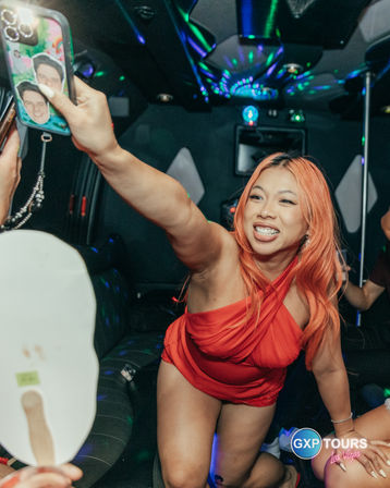 Smiling woman with orange hair in a red dress reaching to take a selfie inside a disco-lit party limo