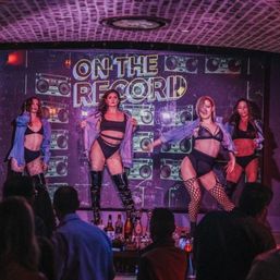 Four dancers in black lingerie, fishnet stockings and thigh-high boots perform a choreographed burlesque-style routine on a nightclub stage beneath an "ON THE RECORD" sign while a bar crowd watches