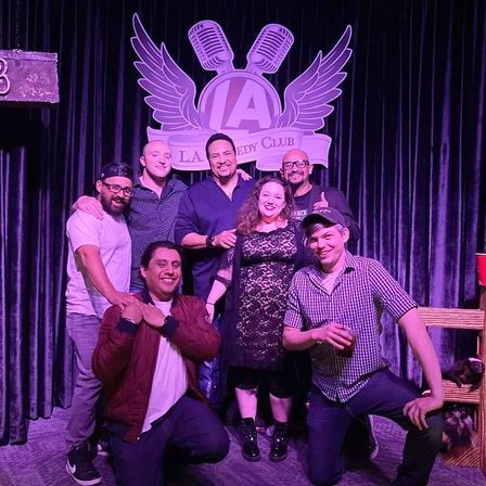 Seven friends posing and smiling on a comedy club stage under purple lighting with a winged microphone logo backdrop, enjoying a night out.