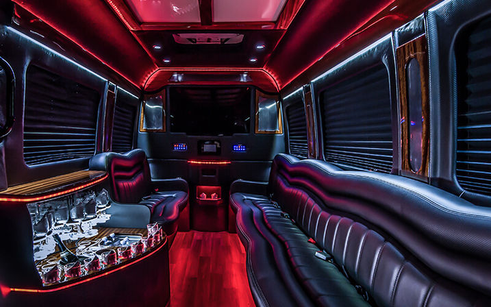 Party-ready luxury stretch limousine interior with curved black leather seating, red and blue LED accent lighting, hardwood floor, stocked mini-bar with glassware and darkened window shades.