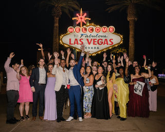 Large celebratory group in formalwear cheering and posing at night in front of the illuminated "Welcome to Fabulous Las Vegas Nevada" sign with palm trees in the background.
