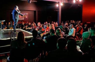 Stand-up comedian on a lit stage performing to a packed, laughing audience in an intimate indoor comedy club under warm spotlights.