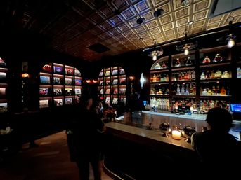 Cozy dimly lit cocktail bar interior with backlit liquor shelves, arched photo-display wall, ornate tin ceiling and a candlelit marble bar with silhouetted patrons