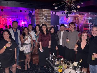 Group of a dozen adults posing in a lively nightclub lounge with purple stage lighting, exposed brick column and retro sputnik chandelier, everyone holding cocktails and champagne around a table with bottles, ice bucket and citrus garnishes, poster-covered walls and stage visible in the background.