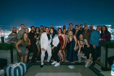 Cheerful group posing for a rooftop night party photo with an illuminated city skyline in the background, dressed in evening attire and holding cocktails