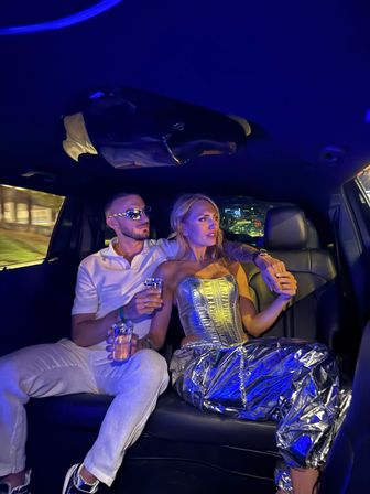 Stylish couple riding a luxury limousine at night with blue ambient lighting — woman in metallic silver corset and pants, man in white shirt and sunglasses holding drinks, city lights visible outside.
