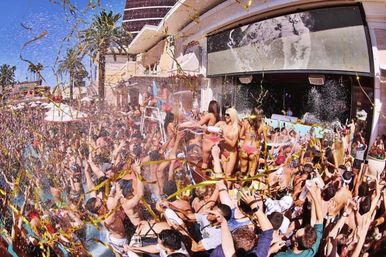 Crowded outdoor pool party at a dayclub with bikini dancers on a raised platform, confetti and gold streamers flying over a sunlit crowd, palm trees and bright blue sky