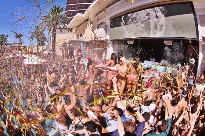 Crowded outdoor pool party at a dayclub with bikini dancers on a raised platform, confetti and gold streamers flying over a sunlit crowd, palm trees and bright blue sky