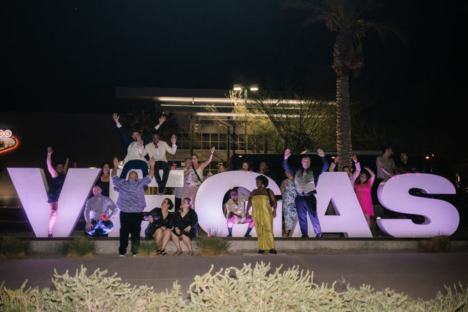 Group of people posing and cheering on giant illuminated "VEGAS" letters at night in Las Vegas, Nevada, with a palm tree and city lights, festive party vibe.