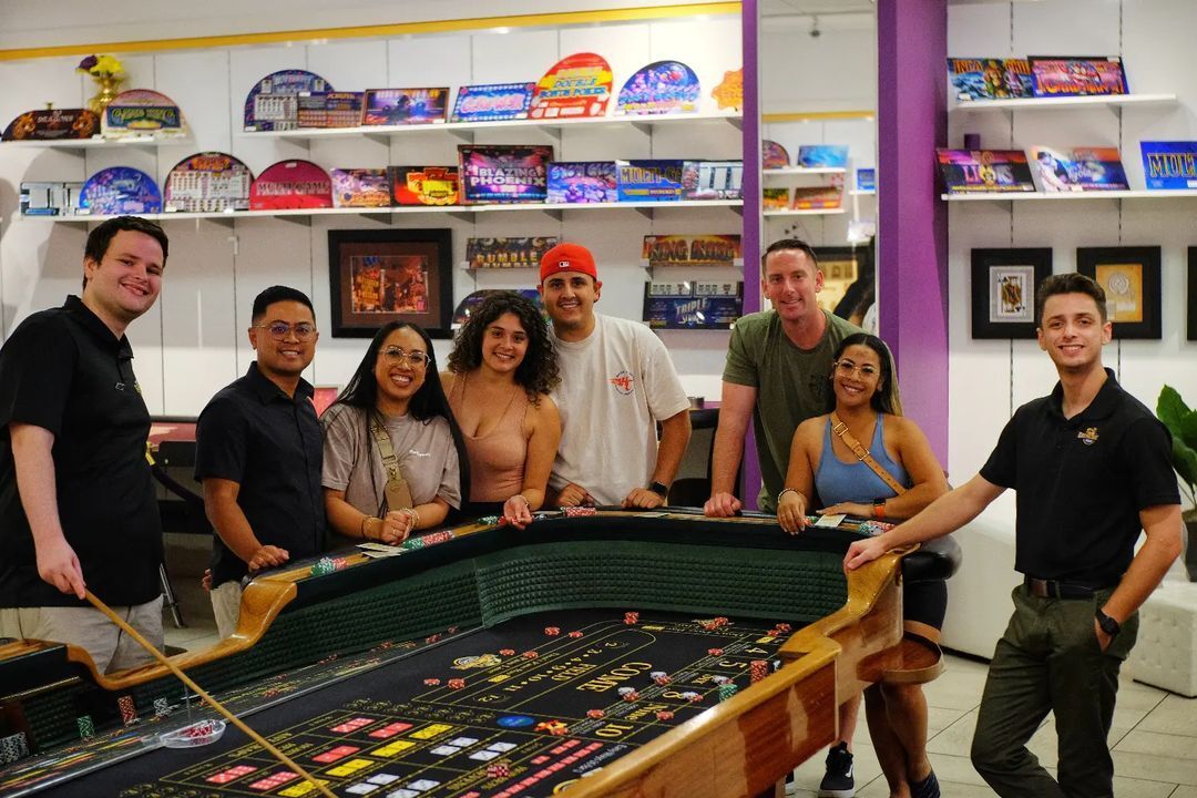 Group of eight people smiling and posing around a casino craps table with chips and stick, colorful game signage and display shelves on an indoor gaming floor.