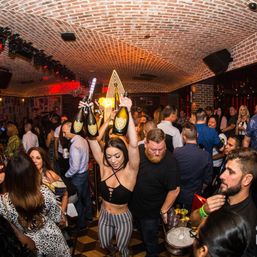 Lively nightclub lounge with arched brick ceiling and crowded dance floor as a server parades multiple champagne bottles overhead for bottle service